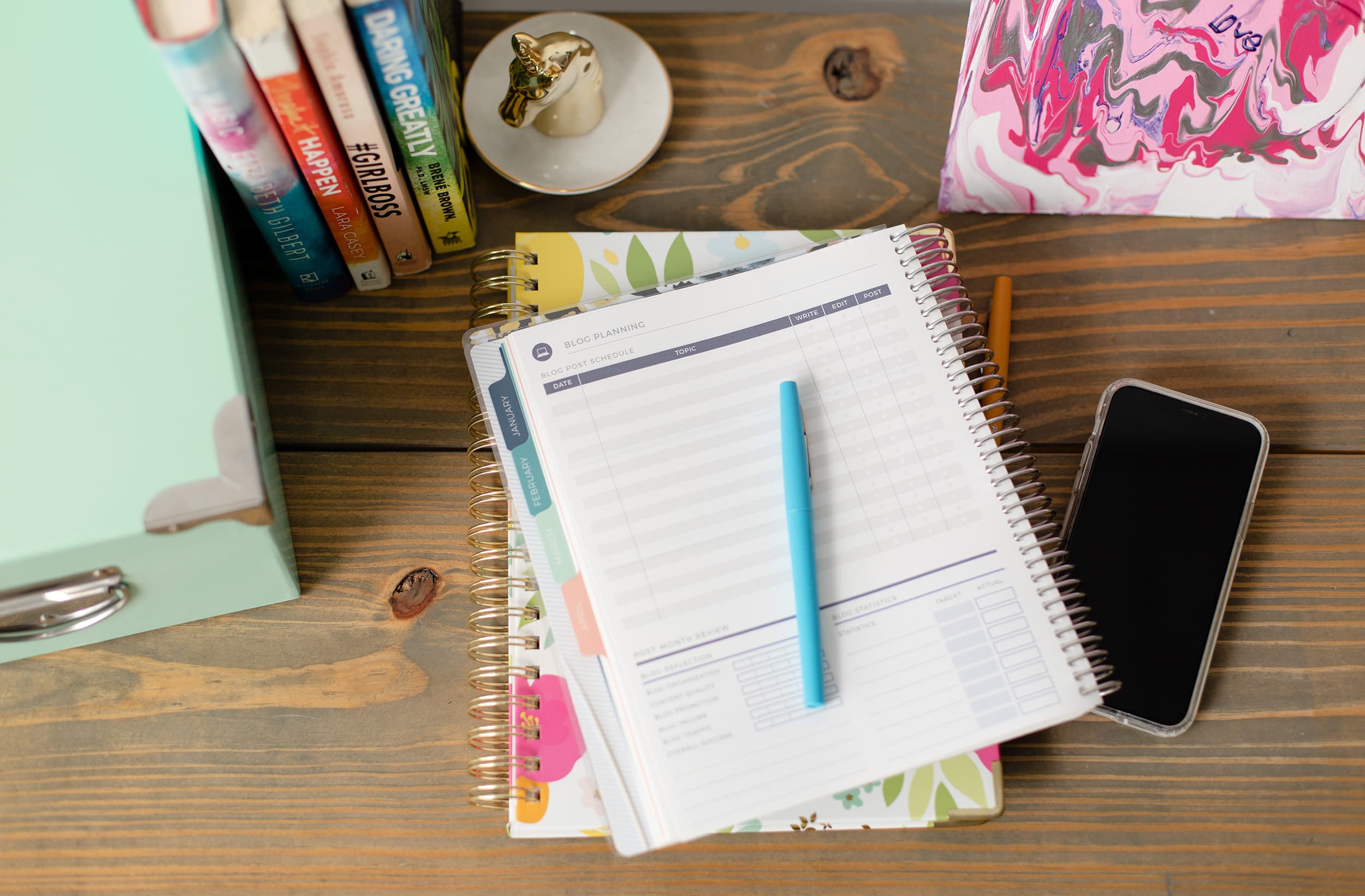 Overhead shot of a desktop with business-related books, a golden unicorn ring dish, two planners, and a cellphone